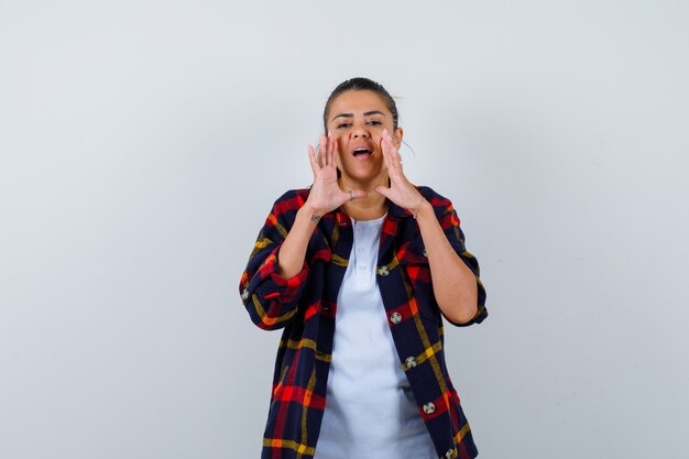 Belle femme en t-shirt blanc, chemise à carreaux avec les mains près de la bouche comme racontant le secret et semblant concentrée, vue de face.