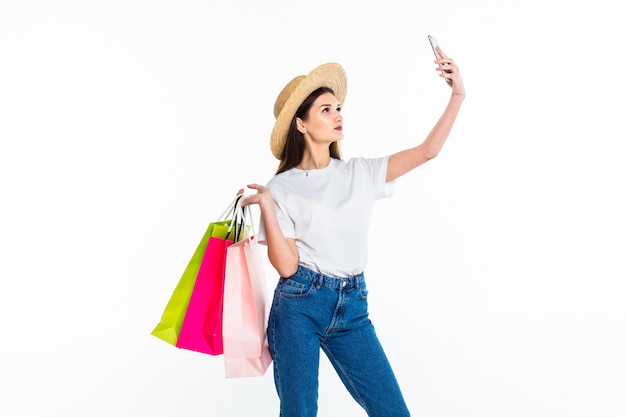 Belle femme tenant des sacs à provisions et prenant selfie avec téléphone portable isolé sur mur blanc