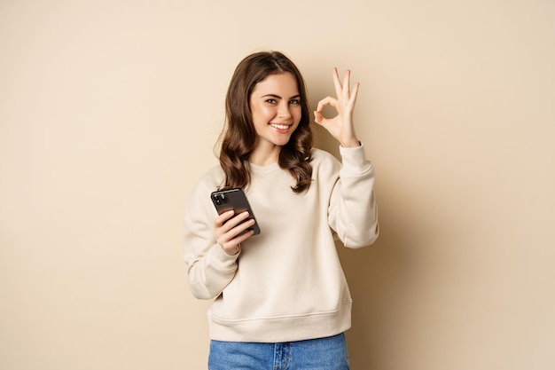 Belle femme tenant un téléphone portable, un téléphone portable et un signe correct, recommandant une application, une application d'achat, debout sur un fond beige.