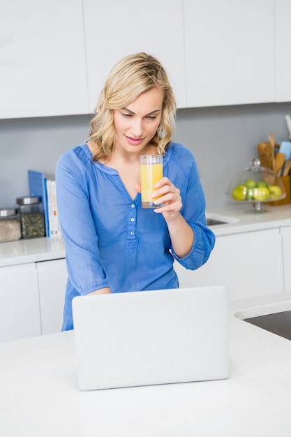 Belle femme tenant un verre de jus utilisant un ordinateur portable dans la cuisine