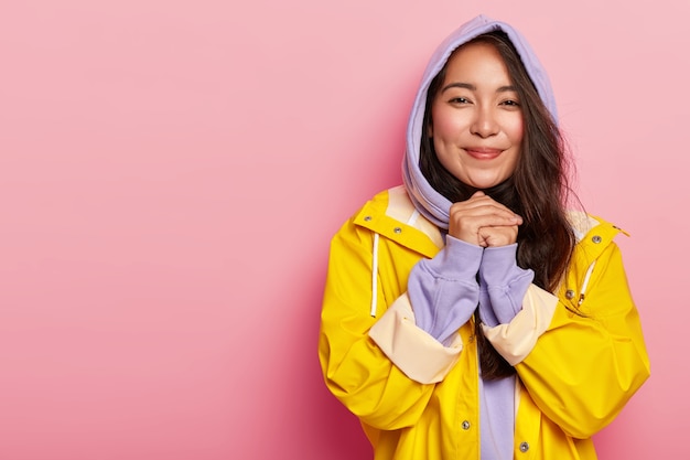 Belle femme touchée avec une apparence agréable, garde les mains jointes, étant touchée par quelque chose, porte un sweat à capuche et un imperméable