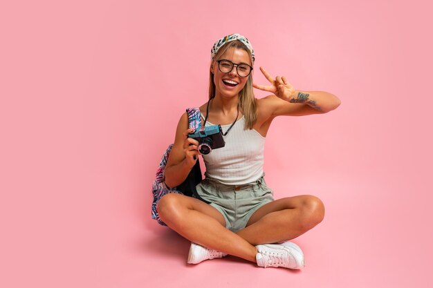 Belle femme touristique avec appareil photo et sac à dos assis sur le sol en studio sur fond rose Ambiance de vacances