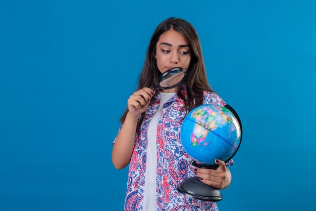 Belle femme touristique tenant et regardant à travers la loupe sur le globe avec intérêt debout sur l'espace bleu