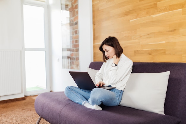 Belle femme travaille sur un ordinateur portable assis sur un lit sombre devant un mur en bois à la maison