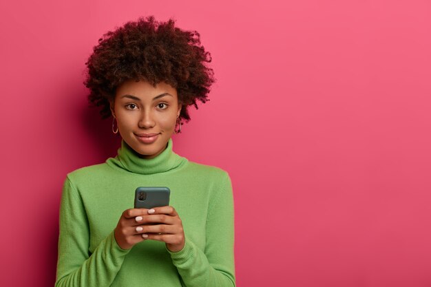 Belle femme avec des types de cheveux afro message sur smartphone, navigue sur le réseau, regarde avec confiance la caméra