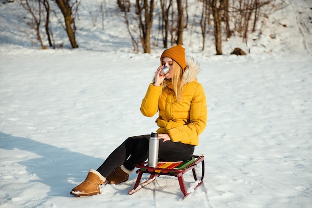 Belle femme en veste d'hiver jaune assise sur le traîneau et buvant du thé chaud du thermos dans le parc enneigé