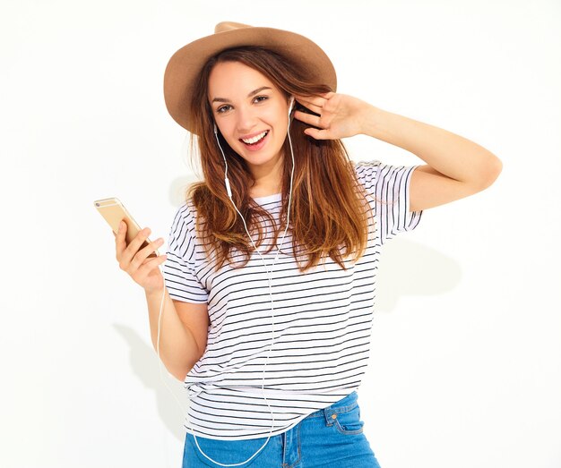 Belle femme en vêtements d'été hipster décontracté sans maquillage en chapeau brun, écouter de la musique au téléphone avec un casque isolé sur blanc