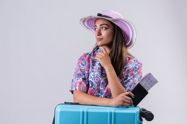 Belle femme de voyageur portant chapeau d'été debout avec valise de voyage tenant des billets à côté avec la main près du menton pensant avoir des doutes