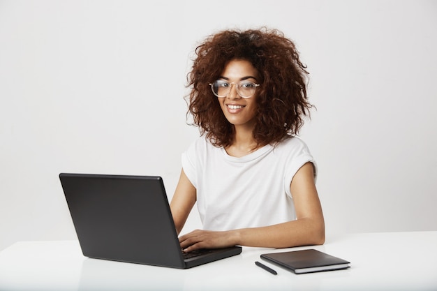 Photo gratuite belle fille africaine souriante assise avec ordinateur portable sur le lieu de travail. mur blanc.