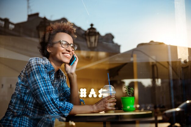 Belle fille africaine souriante, parlant au téléphone, assis dans un café. Tiré de l'extérieur.