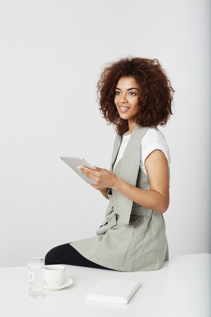 Photo gratuite belle fille africaine souriante tenant la tablette assis sur le lieu de travail. copiez l'espace.