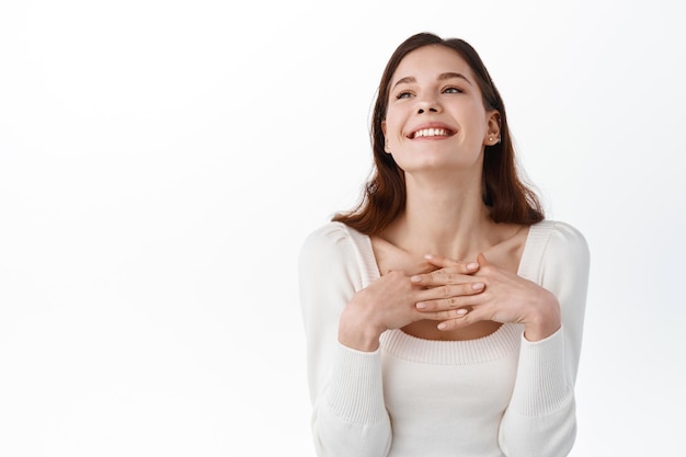 Belle fille à l'air pleine d'espoir, tenant les mains sur la poitrine avec une expression reconnaissante regardant de côté, souriante joyeuse, appréciant les louanges ou les compliments, debout avec les mains sur la poitrine sur fond blanc