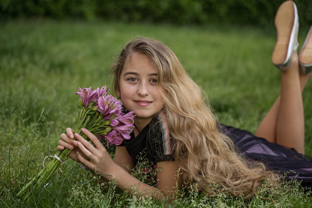 Belle fille allongée sur l'herbe, souriant tout en tenant des fleurs à l'extérieur en t-shirt noir pendant la journée.