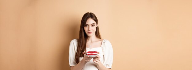 Belle fille d'anniversaire tenant un gâteau avec une bougie faisant un souhait célébrant bday sur fond beige