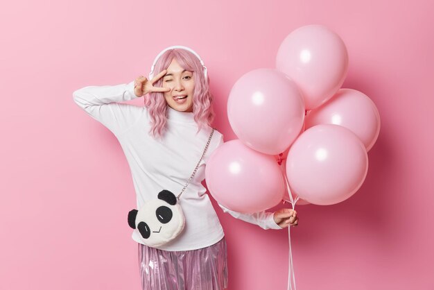 Belle fille asiatique aux cheveux roses fait un geste de paix sur l'œil tire la langue idiote autour de la fête habillée de façon festive tient un tas de ballons gonflés à l'hélium pose pour faire de la photo