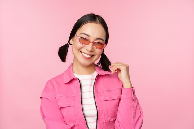 Belle fille asiatique en lunettes de soleil souriant à la caméra posant sur fond de studio rose