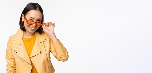 Belle fille asiatique à lunettes de soleil souriant et heureux posant en veste jaune contre blanc b