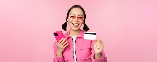 Belle fille asiatique moderne riant et souriant avec une carte de crédit de téléphone portable faisant des achats en ligne en payant avec un smartphone debout sur fond rose