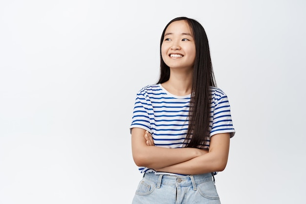 Belle fille asiatique regardant de côté avec un sourire heureux croiser les bras sur la poitrine en regardant quelque chose sur la gauche debout sur fond blanc