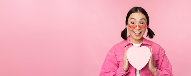 Belle fille asiatique souriante heureuse montrant une boîte-cadeau coeur et regardant excitée à la caméra debout sur fond romantique rose