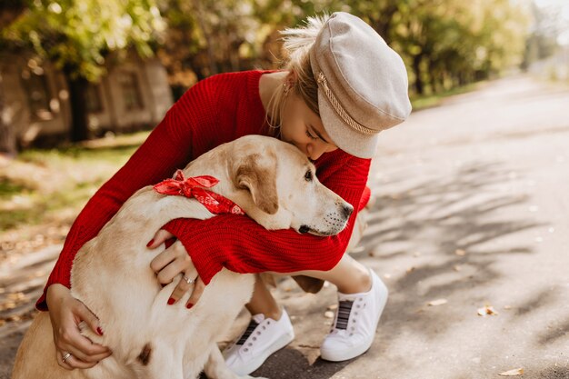 Belle fille au chapeau élégant et baskets blanches tenant tendrement son chien. Belle blonde assise avec son animal de compagnie dans le parc.