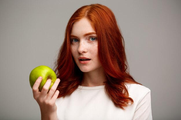 belle fille au gingembre tenant une pomme verte sur le mur gris.