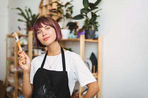 Belle fille aux cheveux colorés en tablier noir et T-shirt blanc tenant un pinceau à la main regardant rêveusement à huis clos au studio de poterie moderne