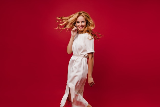 Belle fille aux cheveux longs dansant avec le sourire sur le mur rouge. Heureuse dame caucasienne en robe blanche s'amusant à la fête.