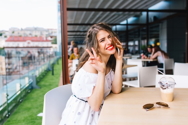 Belle Fille Aux Cheveux Longs Est Assise à Table Sur La Terrasse Du Café. Elle Porte Une Robe Blanche Aux épaules Nues Et Du Rouge à Lèvres Rouge. Elle Parle Au Téléphone Et Sourit à La Caméra.