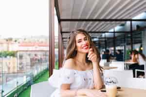 Photo gratuite belle fille aux cheveux longs est assise à table sur la terrasse du café. elle porte une robe blanche aux épaules nues et du rouge à lèvres rouge. elle sourit à la caméra .