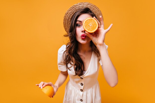 Belle fille aux cheveux noirs aux yeux verts en chapeau et robe d'été envoie un baiser et pose avec des moitiés orange.