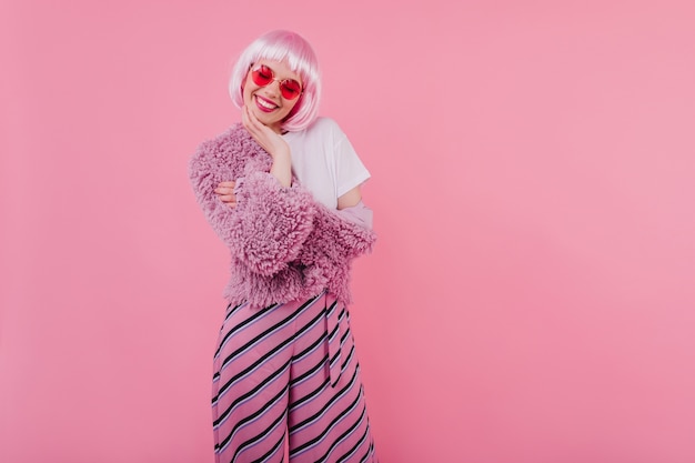 Belle fille aux cheveux roses courts riant les yeux fermés. jeune femme raffinée porte peruke et lunettes à la mode pendant la séance photo