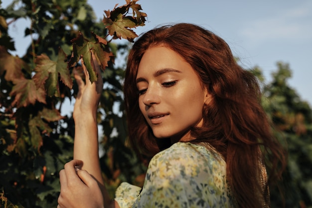 Belle fille aux cheveux rouges avec des taches de rousseur en tenue cool jaune et verte regardant vers le bas et posant sur les vignes