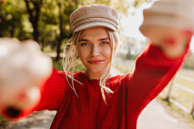 Belle fille blonde dans un joli chapeau à la mode et un pull rouge faisant selfie dans le parc en automne.