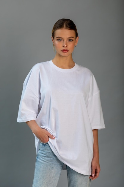 Une belle fille blonde dans un t-shirt blanc surdimensionné et un jean bleu posant sur un fond gris