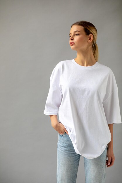 Une belle fille blonde dans un t-shirt blanc surdimensionné et un jean bleu posant sur un fond gris
