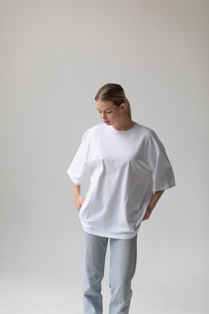 Photo gratuite une belle fille blonde dans un t-shirt blanc surdimensionné et un jean bleu posant sur un fond gris