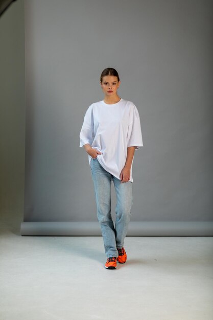 Une belle fille blonde dans un t-shirt blanc surdimensionné et un jean bleu posant sur un fond gris