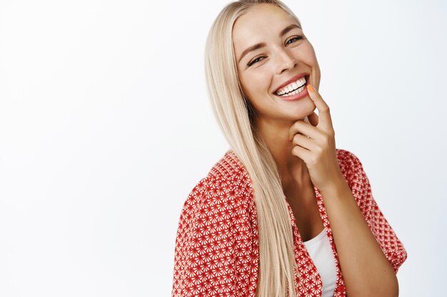 Belle fille blonde avec de longs cheveux sains riant et souriant à la caméra debout dans des vêtements d'été sur fond blanc