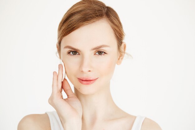 Belle fille blonde naturelle, nettoyer le visage avec une éponge de coton en souriant. Cosmétologie et spa.