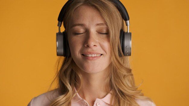 Belle fille blonde rêveuse écoutant de la musique dans un casque avec les yeux fermés sur fond jaune