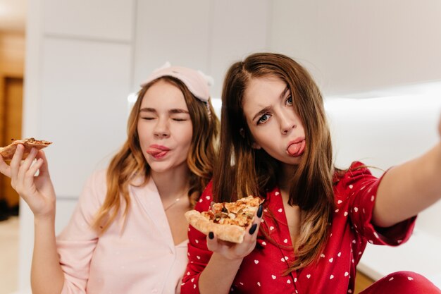 Belle fille bouclée en masque pour les yeux rose tenant une tranche de pizza. Femme aux cheveux noirs en tenue rouge faisant selfie pendant le petit déjeuner.