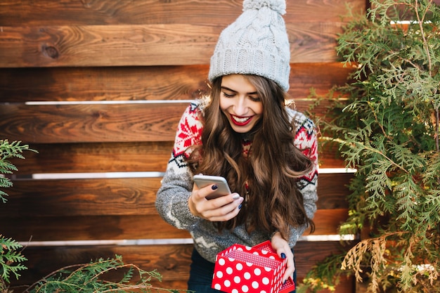 Belle fille brune aux cheveux longs et aux lèvres rouges sur l'extérieur en bois. Elle porte un bonnet tricoté, un téléphone et une boîte à cadeaux. Elle a l'air heureuse.