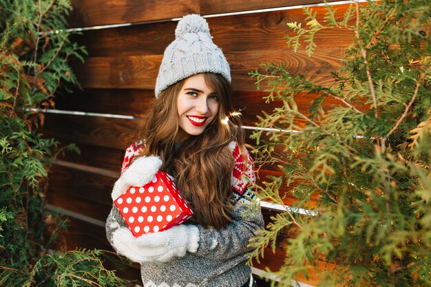 Belle fille brune aux cheveux longs en vêtements d'hiver avec cadeau de Noël sur l'extérieur en bois. Elle sourit.