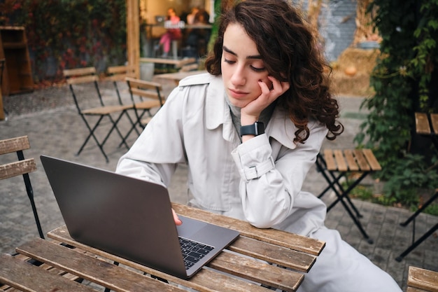 Photo gratuite belle fille brune décontractée en trench-coat travaillant pensivement sur un ordinateur portable dans un café en plein air