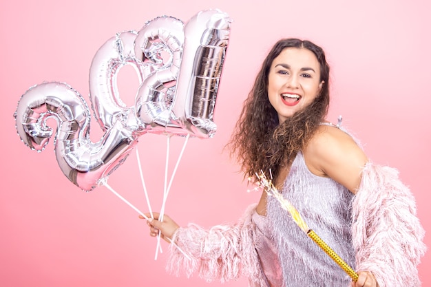 Belle fille brune joyeuse habillée de façon festive aux cheveux bouclés sur un fond de studio rose posant avec une bougie de feux d'artifice à la main et des ballons d'argent pour le concept de nouvel an