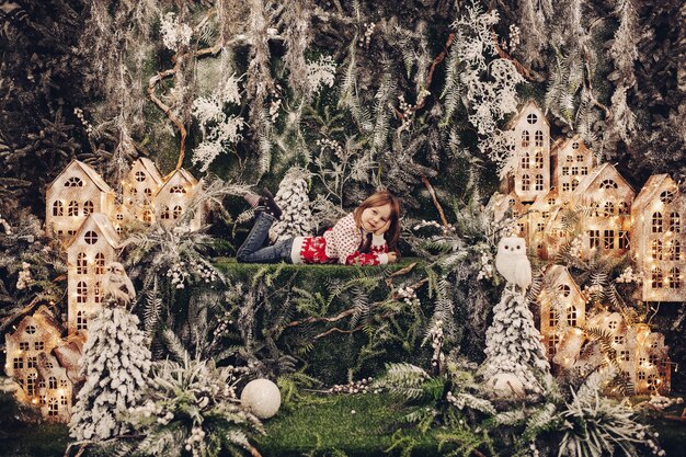 Belle fille brune en pull d'hiver et jeans portant sur une pelouse verte entourée de maisons illuminées faites à la main et de branches de sapin recouvertes de neige artificielle. Conte de Noël.