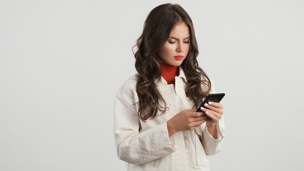 Belle fille brune sérieuse utilisant attentivement un smartphone sur fond blanc