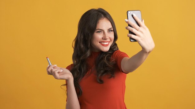 Belle fille brune souriante avec du rouge à lèvres prenant joyeusement selfie sur smartphone sur fond coloré