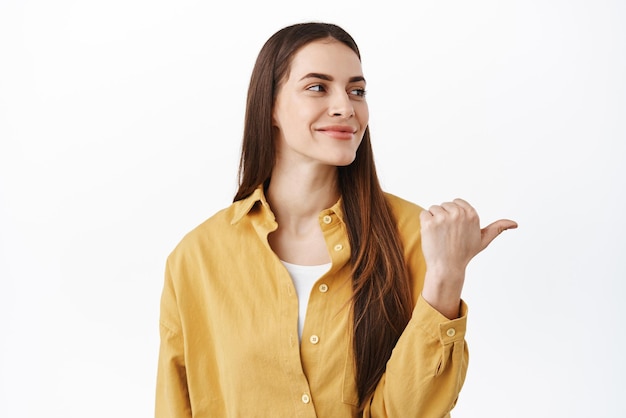 Belle fille candide aux cheveux longs et à la peau naturelle tourne la tête et pointe de côté vers l'espace de copie droit souriant heureux confiant sur le choix debout sur fond blanc